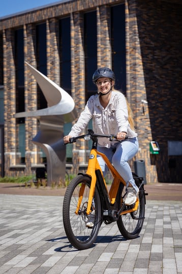 Vrouw met helm op een oranje speed pedelec, fietsend.