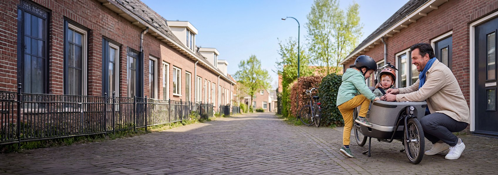 Kindje stapt bij vader in de bakfiets, terwijl vader en ander kindje wachten.