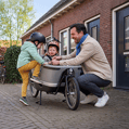 Vader zet kindje vast in zijn bakfiets terwijl het andere kindje zelf in de elektrische bakfiets klimt.