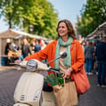 Vrouw loopt met haar scooter bij de markt weg met een tas boodschappen in haar hand.