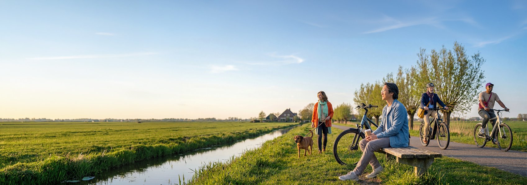 Een mevrouw laat de hond uit langs een polder fietspad, waar iemand op een bankje zit en waar 2 mensen langsfietsen.