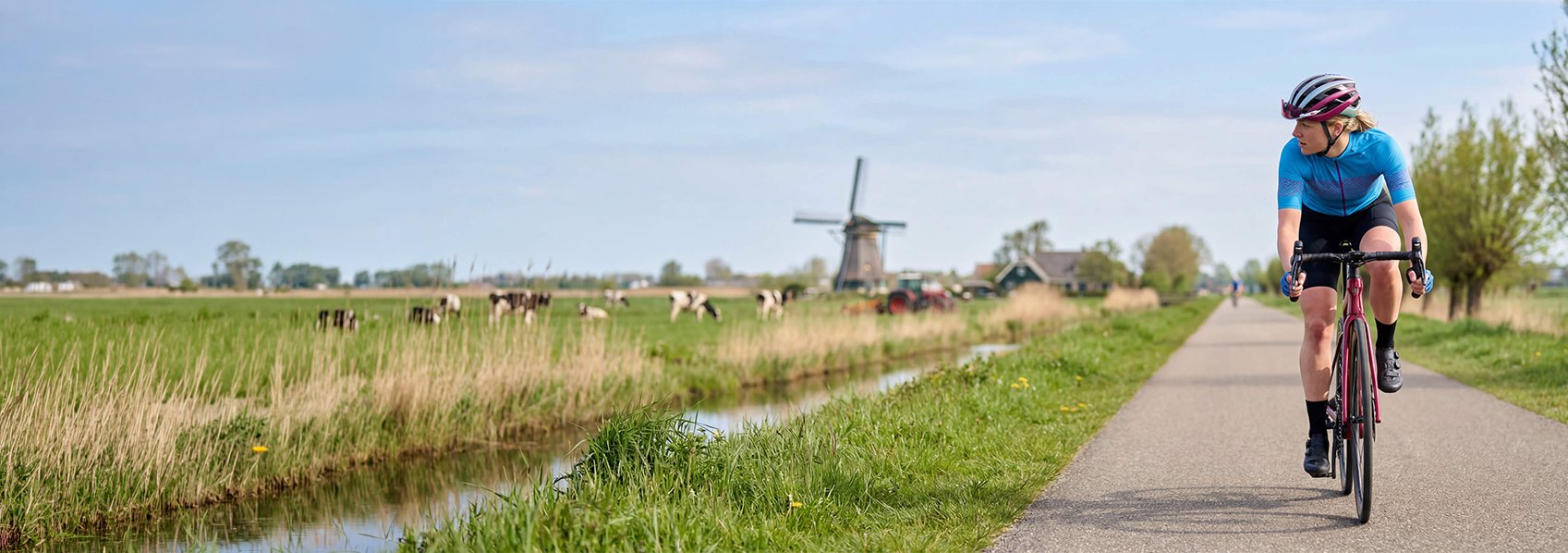 Op een polder fietspad met koeien en een molen fietst een blonde wielrenster op een racefiets.