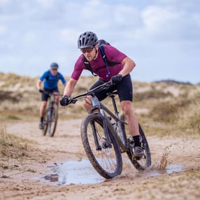 Twee mountainbikers mountainbiken door de duinen, door zand en plassen.