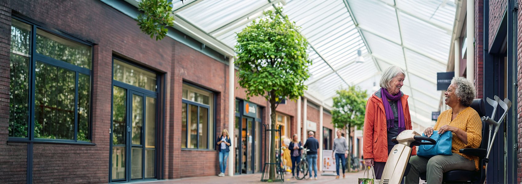 Dame in een scootmobiel staat met een andere oudere dame te praten in een winkelcentrum.